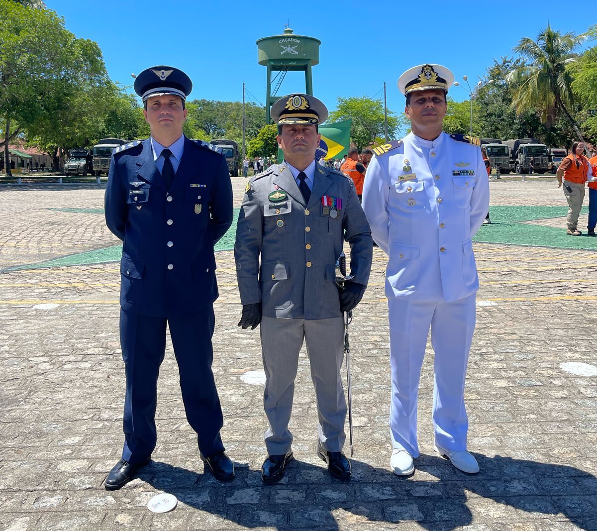 Três militares em uniformes diferentes, em cerimônia ao ar livre.