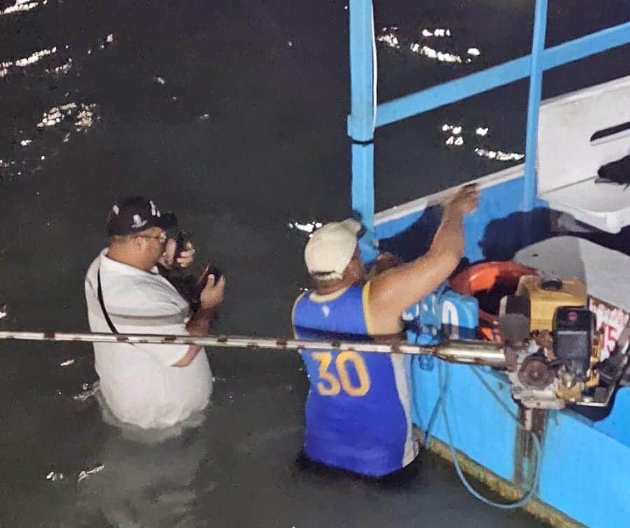 Homem fotografa barco à noite na água rasa.