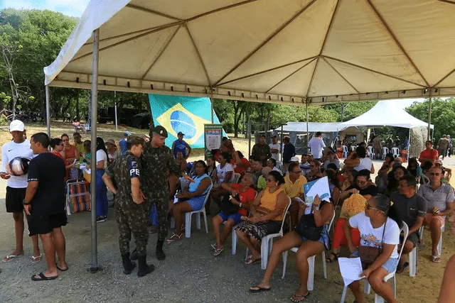 Pessoas aguardam em fila sob tenda com bandeira.