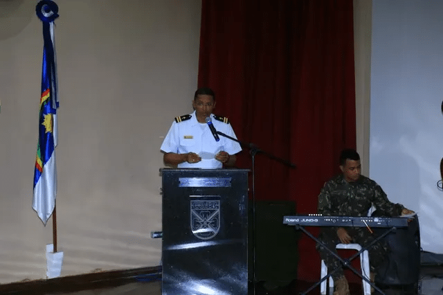 Oficial discursa em evento com pianista ao lado.