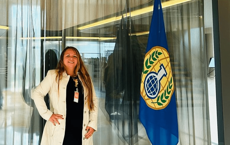 Mulher de pé ao lado de bandeira institucional.