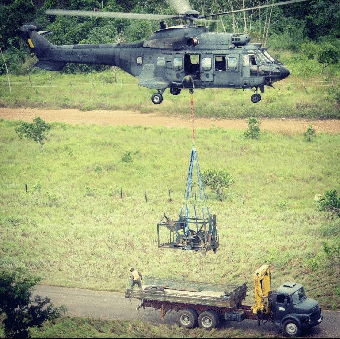 Helicóptero transporta equipamento sobre caminhão em área rural.