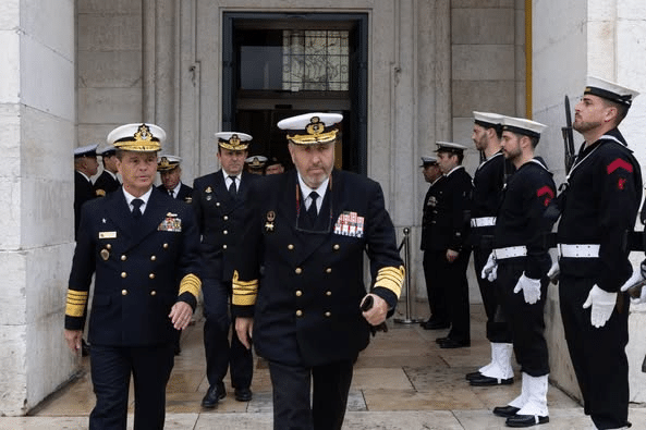 Oficiais navais uniformizados saindo de prédio oficial.