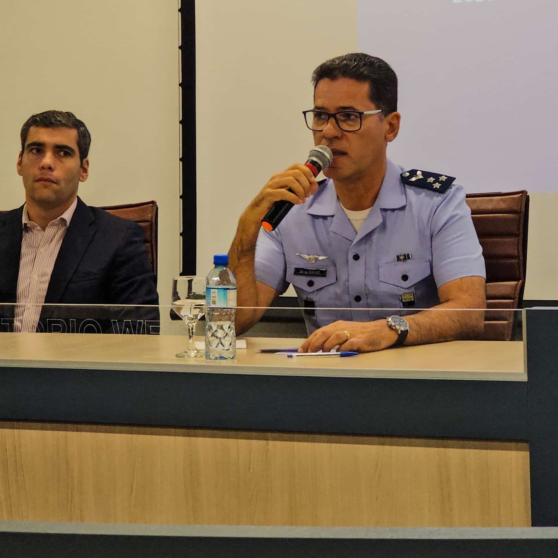 Oficial falando em conferência, microfone na mão, uniforme azul.