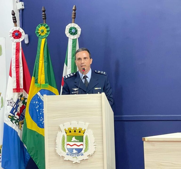 Oficial discursa em púlpito com bandeiras ao fundo.