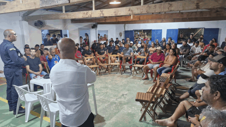 Reunião comunitária em espaço coberto, pessoas ouvindo palestrantes.