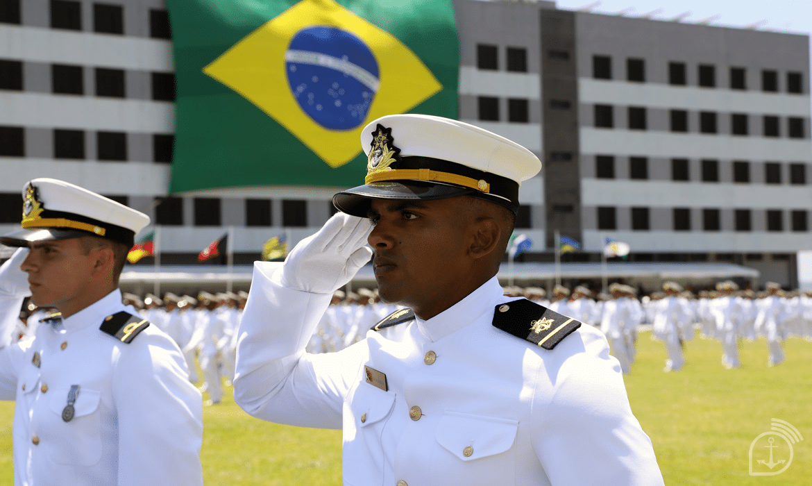 Militares em uniforme branco, bandeira do Brasil ao fundo.