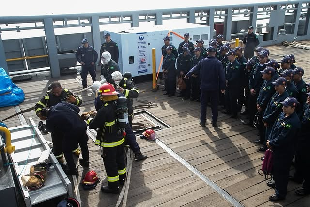 Equipe de bombeiros e trabalhadores em plataforma portuária.