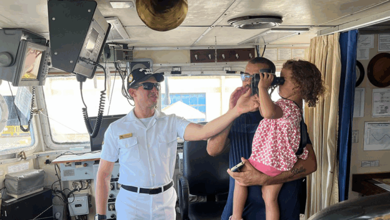 Criança usa binóculo em navio com marinheiros.