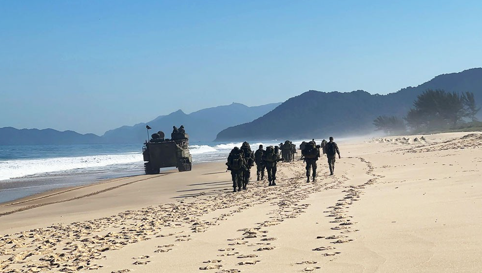Soldados marchando na praia com veículo militar.