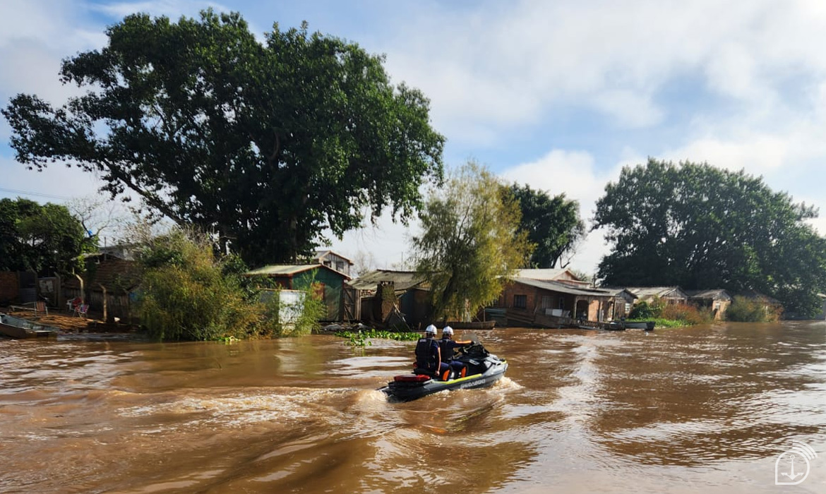 Casa alagada próxima a rio, barco de resgate passando.