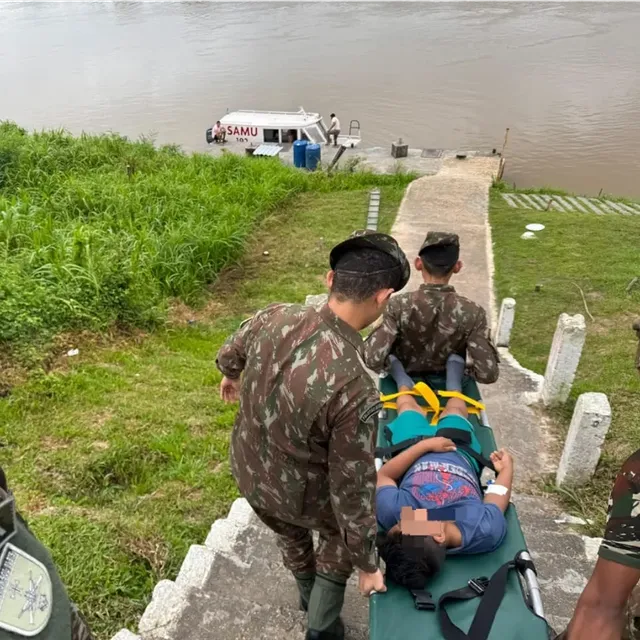 Soldados carregam maca perto de ambulância e rio.
