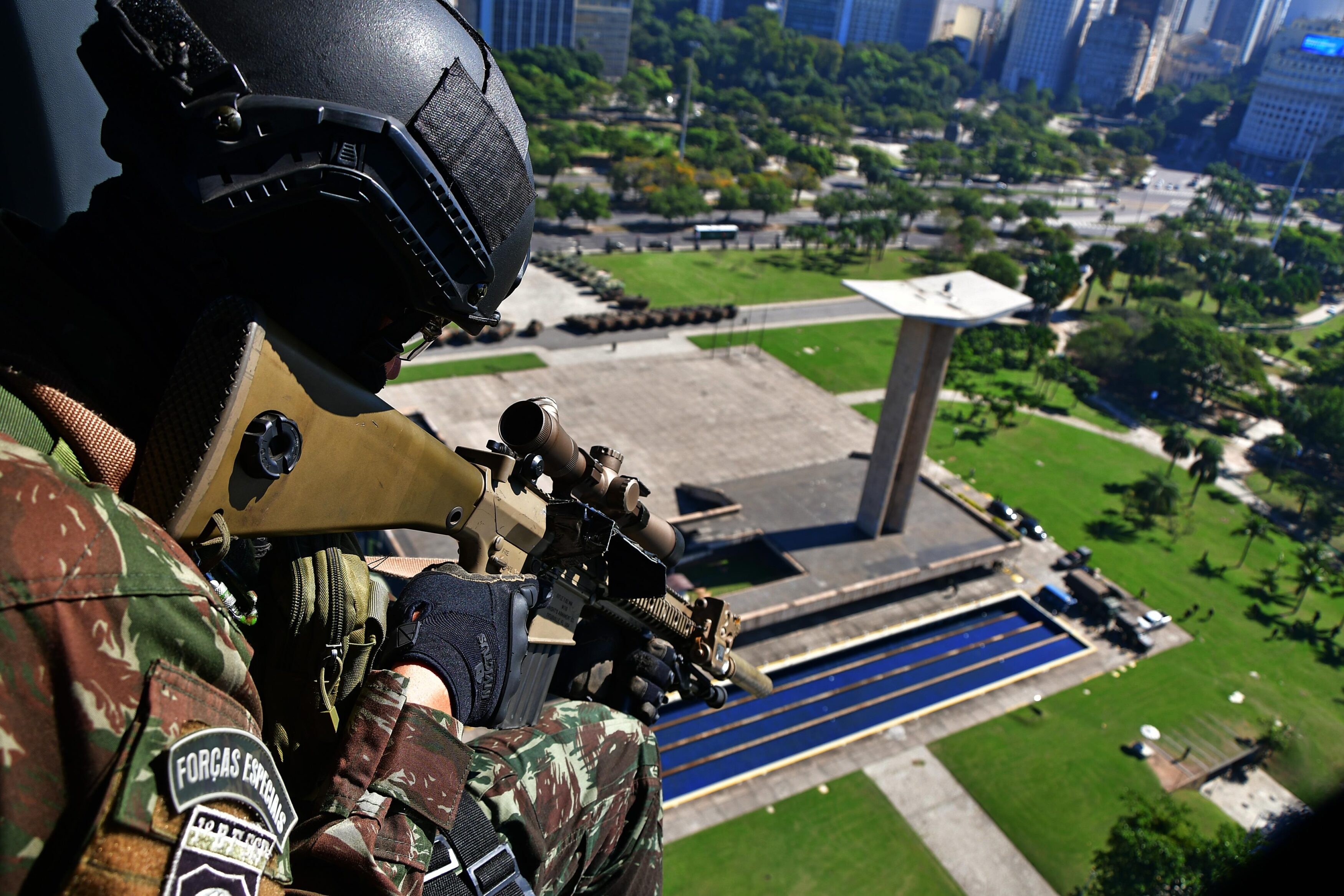 Soldado armado em helicóptero sobre cidade brasileira, monumento ao fundo.