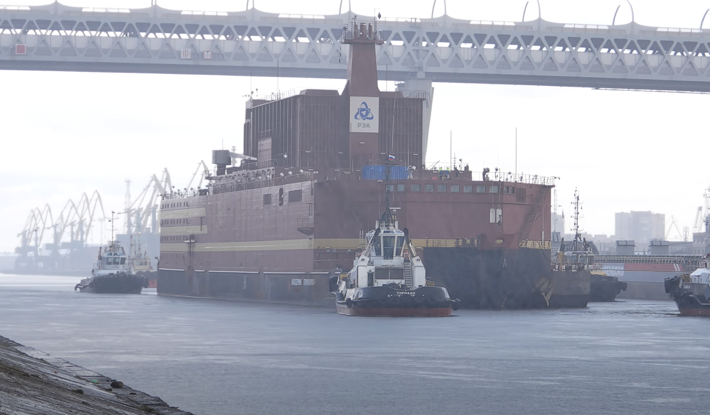 Rebocador transportando plataforma nuclear sob ponte.