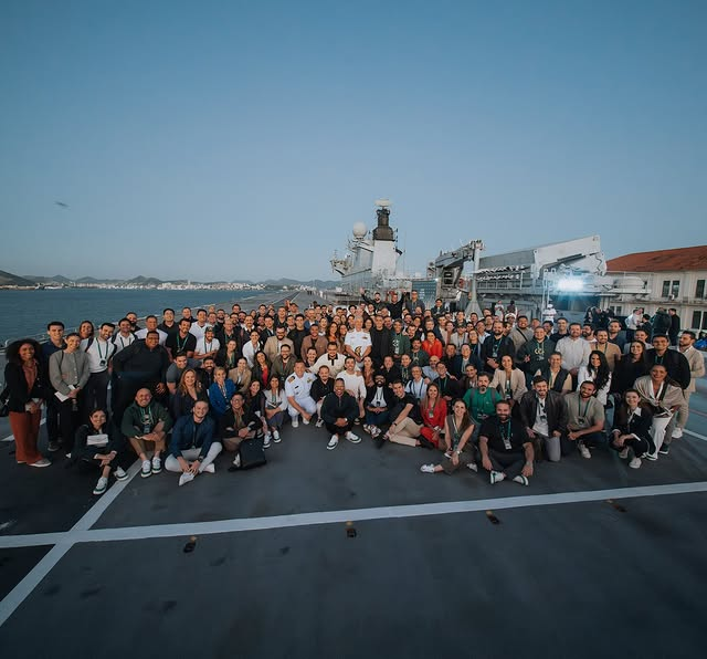 Grupo de pessoas em convés com navio ao fundo.