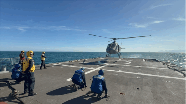 Helicóptero pousando em navio com equipe de apoio.