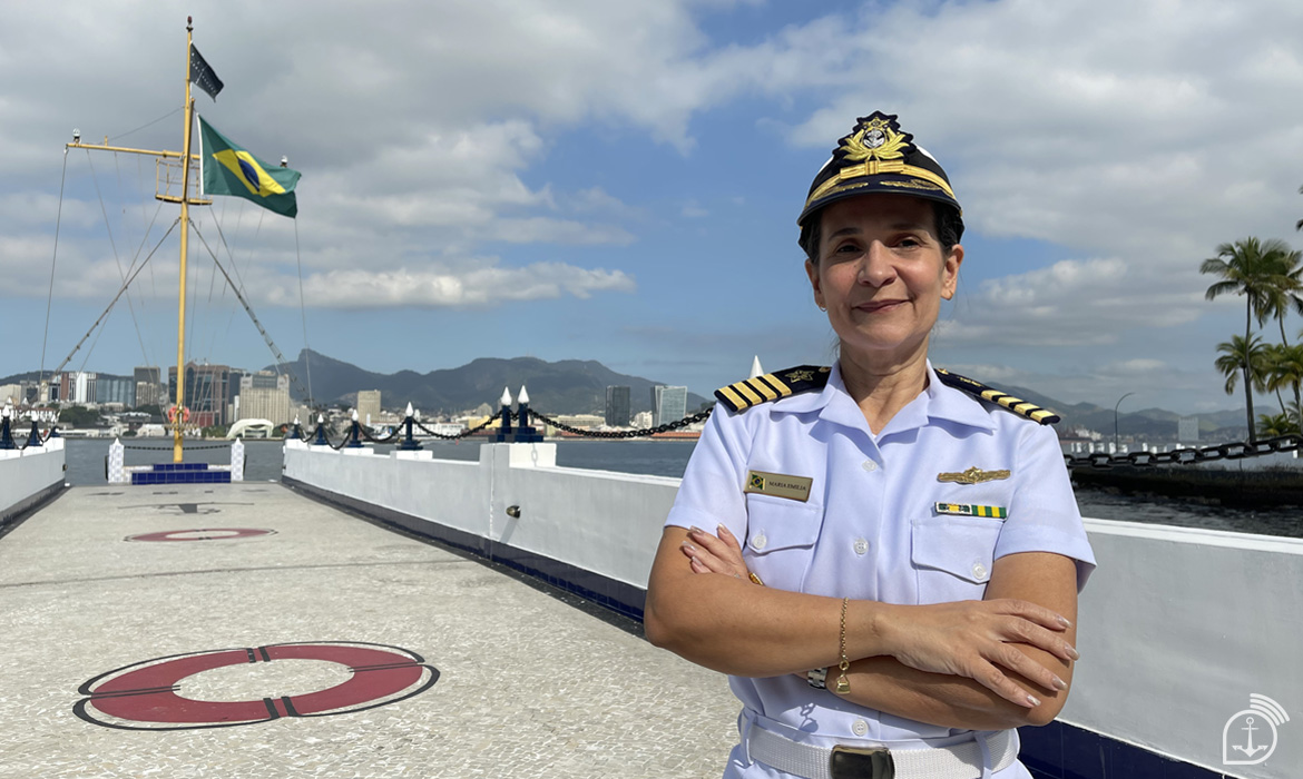 Oficial naval em uniforme, bandeira do Brasil ao fundo.